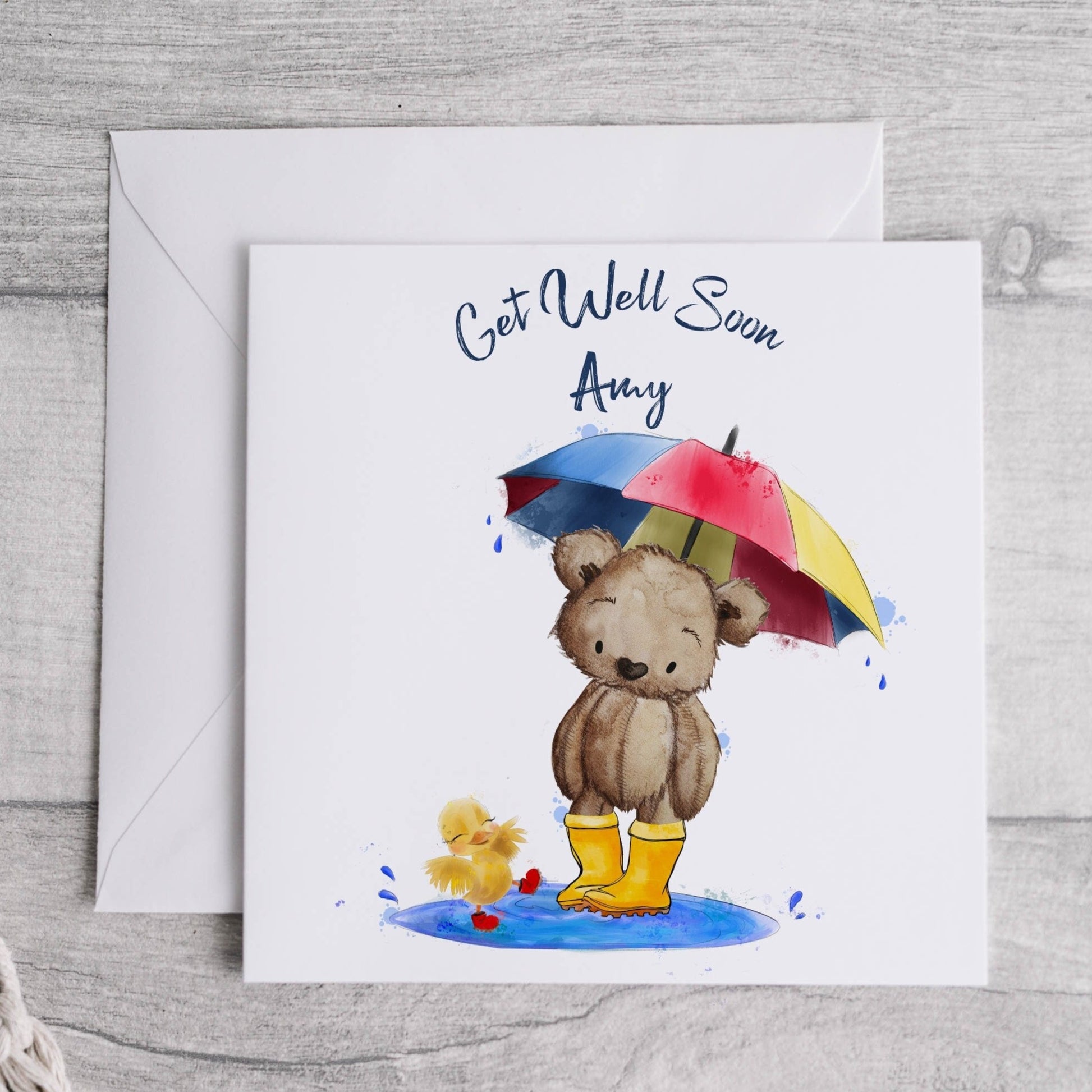 Square card with an image of a teddy bear in wellies holding an umbrella with a chick splashing in a puddle. Text reads 'Get well soon' and is personalised with a name